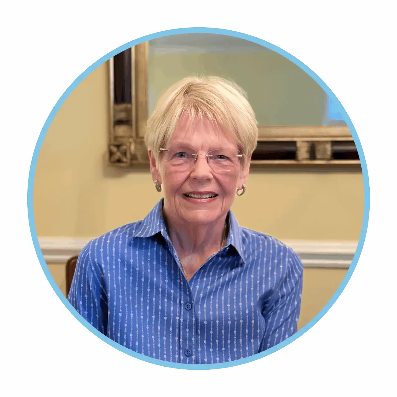 An older woman with short blonde hair, wearing glasses and a blue button-up shirt with white stripes, smiles while sitting indoors in front of a beige wall and a framed mirror.